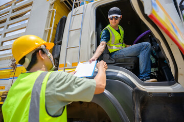 A logistics worker in safety gear hands over documents to a truck driver from outside the vehicle,...