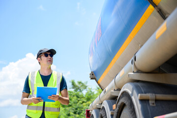 A logistics worker in a reflective vest holds a clipboard as he inspects a large fuel tanker on a...