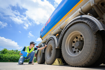 A safety inspector in a reflective vest checks the tires of a large fuel tanker under a bright blue sky. Holding a clipboard, he ensures compliance with transportation safety standards. © เลิศลักษณ์ ทิพชัย