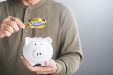 A man holds a white piggy bank and uses a magnifying glass to examine it, symbolizing the careful study of savings, financial planning, and investment strategies for future growth