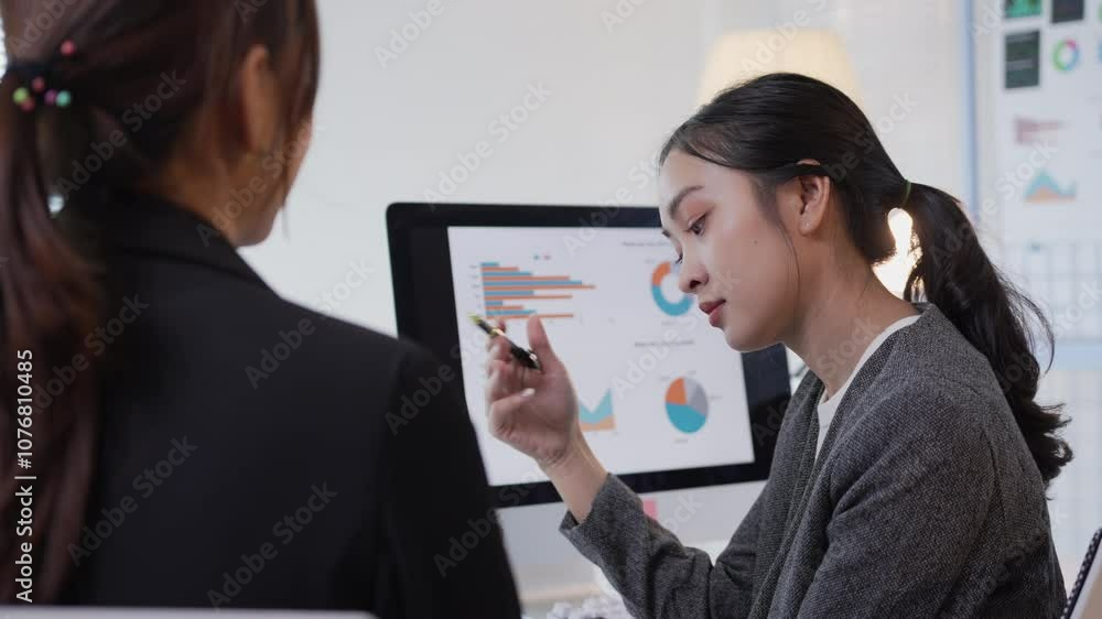 Poster two asian businesswomen discussing charts on a computer while collaborating in a modern office setti