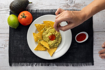 A white plate of nachos with creamy guacamole is surrounded by an avocado, lime, fresh tomato,...