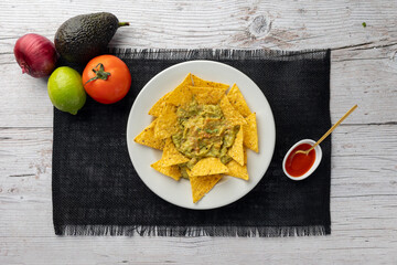 A white plate of nachos with creamy guacamole is surrounded by an avocado, lime, fresh tomato,...