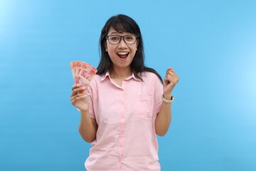 Enthusiastic young asian woman looking excited at camera, holding indonesia money banknotes rupiah in hand, standing over blue background