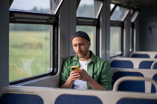 Relaxed man wearing wireless earphones, using smartphone and surfing Internet, social media in train car. Tech enhanced journey, tourist commuting experience, public transit with music and podcasts
