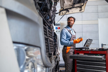 Professional repairman standing by broken vehicle and servicing truck inside workshop.