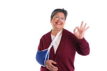 An elderly woman with a smile and arm in a sling making an OK gesture, isolated on a white...