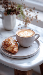 Bright, high-resolution image in cozy cafe setting. Tabletop holds rustic ceramic cup of Cappuccino with latte art beside fresh pastry on plate. Warm lighting and aroma creates tranquil inviting mood