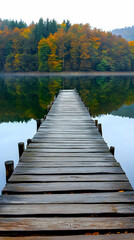 Naklejka premium Wooden pier extending into a still lake with autumn trees in the background.