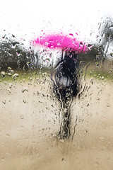Person holding a pink umbrella protecting themselves from the rain, blurred, seen through a wet glass, with drops and condensation. Water, winter, autumn, getting wet and raining.
