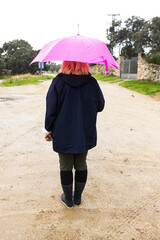 Medium shot of a woman from behind dressed in a blue raincoat and holding a pink umbrella, covering herself from the rain. Water, winter, autumn, getting wet and raining.