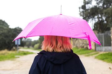 Close-up shot of a woman from behind wearing a blue raincoat and holding a pink umbrella, covering herself from the rain. Water, winter, autumn, getting wet and raining.