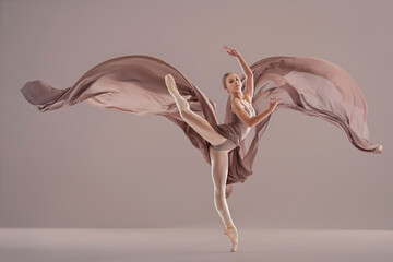 Young ballerina girl in pointe shoes dancing with developing beige fabric on uniform background