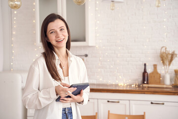 Smiling woman using tablet in cozy kitchen with warm lighting and string lights.