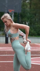 Sporty happy young blonde woman in sportswear doing leg stretching exercises standing on basketball...