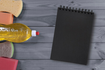 Blank notepad with cleaning supplies on gray wooden background suggesting housekeeping checklist or planning concept. Black page with empty space for copyspace, tips, hacks, motivational text