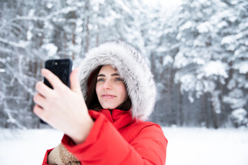 Beautiful brunette in winter in forest in red jacket with hood on her head with smartphone in hand.