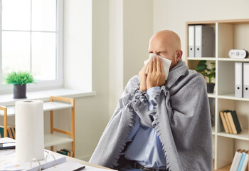Sick man suffering from runny nose sneezing in a napkin sitting on his workplace in office. Male...