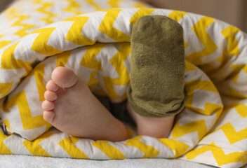The child's feet with one sock stick out from under the blanket. Daytime or morning sleep. Colored blanket.  Happy childhood concept.