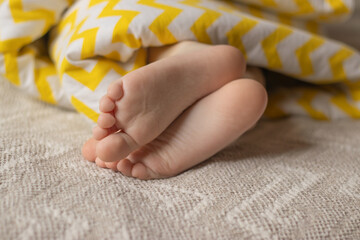 The child's legs stick out from under the blanket.
Baby's bare feet. Daytime or morning nap. Colored blanket.  Happy childhood concept.