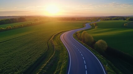 A scenic, winding asphalt road gracefully stretches through vibrant green fields, under a stunningly beautiful sunset sky, creating a perfect backdrop for an enjoyable travel experience