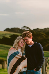 Young Caucasian couple wrapped in a blanket smiling outdoors in a natural space, at sunset