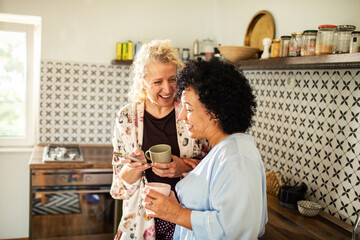 Mature lesbian couple laughing at smartphone in the kitchen with coffee