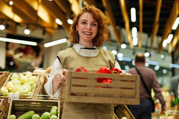 Smiling greengrocer holding crate of red bell peppers in supermarket