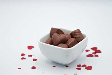 White Dish Full Of Chocolates Surrounded By Red Heart Confetti And Confetti Gems On White Background