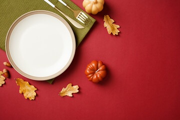 Elegant autumn table setting with white plate, gold cutlery, pumpkins, and oak leaves on a red background. Ideal for fall, Thanksgiving, and seasonal content