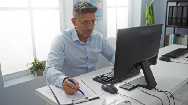 Middle-aged man taking notes at his desk in an office environment, working on a computer with focus and concentration