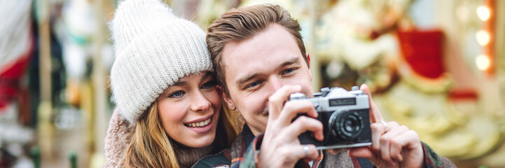 BBeautiful young loving couple, boyfriend and girlfriend having fun on a Christmas market wearing warm clothes, taking pictures on old retro camera. Outdoors, winter time, snowy weather. Banner