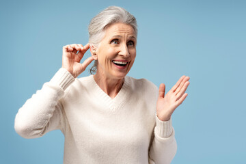 Happy, attractive senior woman having hearing aid in ear, posing isolated on blue background
