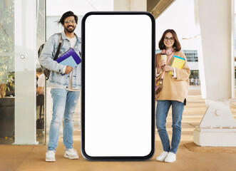 Two young adults stand outdoors beside a large blank smartphone display. One holds a coffee cup, while the other carries colorful notebooks, both smiling in a modern environment. © Prostock-studio