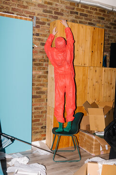 Person in Protective Suit Standing on Chair Reaching High Shelve