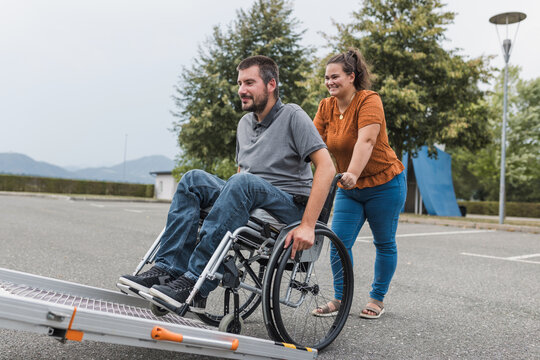Smiling woman, a personal caregiver assisting a man with a disability in entering the adaptive vehicle with an accessible ramp.