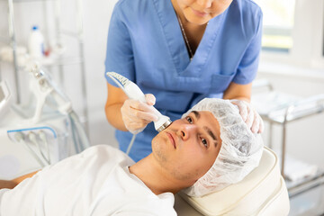 Fototapeta premium Young woman cosmetologist performs facial ultrasound procedure with apparatus young male patient