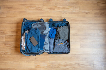 Open suitcase with clothes in it and a smartphone ready to go on a trip. Messy arranged, on a wooden floor in the house.
