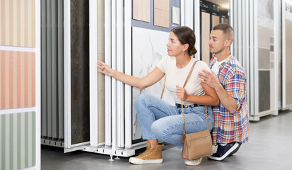 Interested young couple selecting porcelain tiles for home renovation in building hypermarket, carefully examining and discussing options presented on sliding display panels..