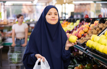 In supermarket,middle-aged woman in traditional Muslim hijab takes and puts ripe apples in small bag