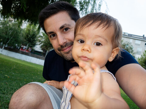 Happy baby boy waving while sitting on father's lap at the park - Powered by Adobe