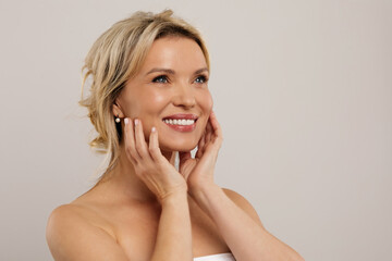 Closeup of middle aged blonde woman isolated on grey background, smiling beautiful naked adult lady touching young looking skin, looking at copy space for ad, studio shot. Injection cosmetology. High © Anastasiya