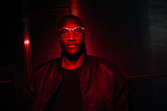 Man illuminated by red light wearing glasses against dark striped back