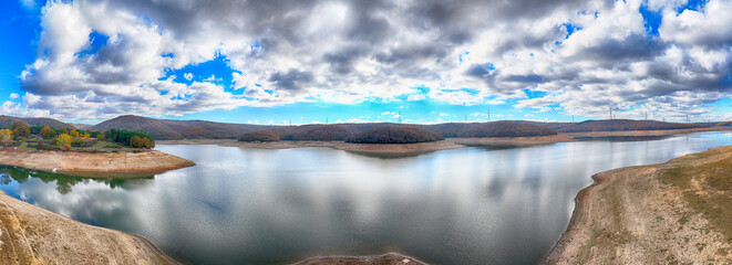 Serene lake nestled amidst rolling hills under a dramatic sky. Aerial panoramic view of a tranquil lake surrounded by tree-covered hills. Drone panorama photography 