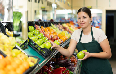 Young girl seller has chosen ripe fruits and holds three shiny sweet apples in hands.Kiwis and apples in boxes in background