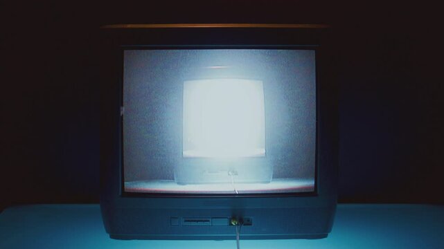 Old television showing brightly glowing TV on its own screen, creating recursive effect in dark studio. Close-up view