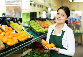 Young woman seller in apron lays out tangerines in supermarket