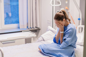 young woman in a blue hospital gown sits alone on a bed in a private room in a clinic, expressing...