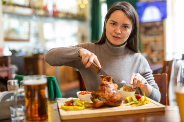 Smiling woman tourist enjoying traditional baked pork knuckle served with pickles, sauces and glass of beer in Viennese restaurant