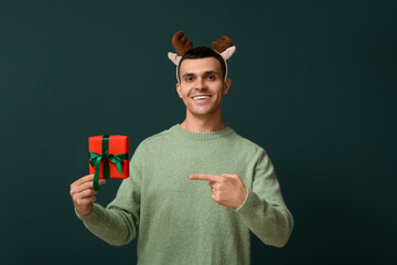 Happy young man in reindeer horns with Christmas gift box on green background